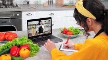 Woman in kitchen watch online webinar laptop listen chef, writes cooking recipe