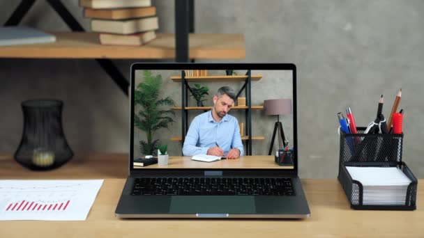 Laptop staat op tafel display met zakenman in kantoor schrijft in notebook — Stockvideo