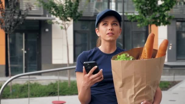 Consegna cibo donna corriere utilizza smartphone tiene sacchetto di carta alla ricerca indirizzo — Video Stock