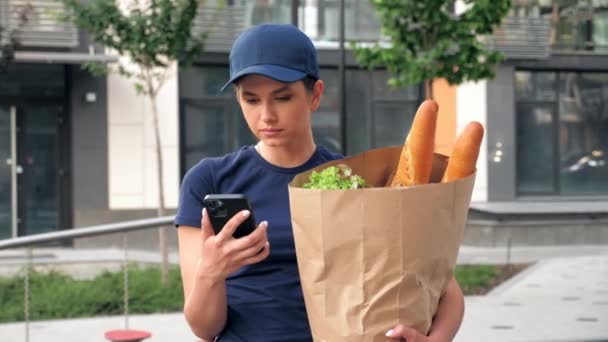 Portrait food delivery woman courier uses smartphone holds paper bag in hand — Stock Video