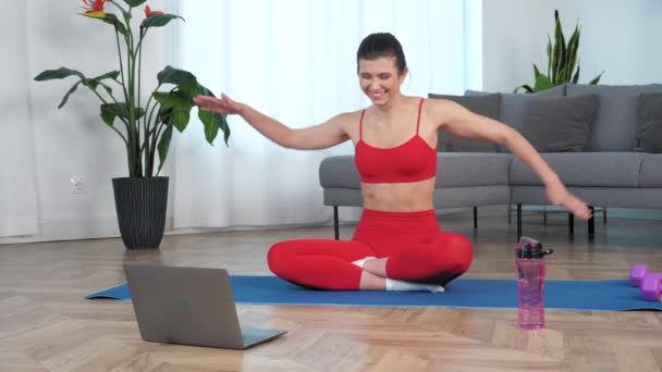 Funny sportive smiling woman sitting on yoga mat makes wave with hands at home — Stockvideo