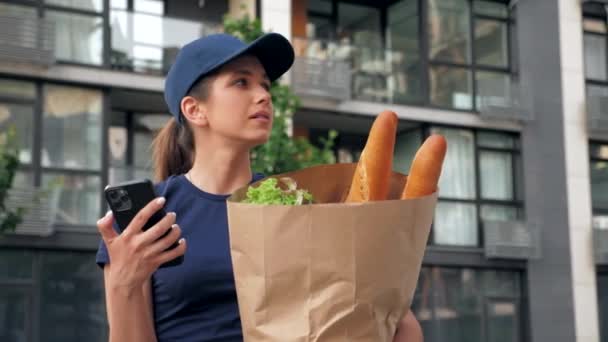 Food delivery woman courier uses smartphone holds paper bag looking address — Stock Video