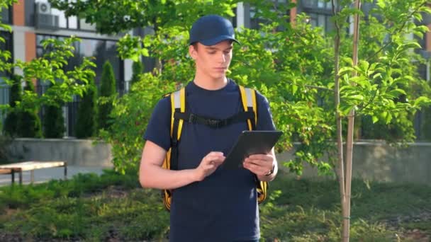 Ampliar cámara en mensajero joven de reparto de comida con mochila térmica utiliza tablet — Vídeo de stock