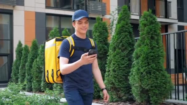 Gülümseyen yemek teslimatçısı kurye caddede yürüyüp akıllı telefondan navigasyonu izliyor. — Stok video