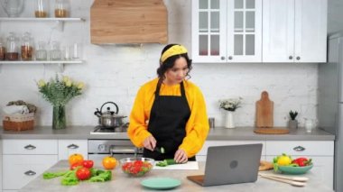 Mutfaktaki kadın salatalık dilimliyor. Şef çevrimiçi yemek kursu laptopu izliyor.