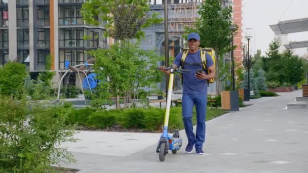 Homem afro-americano mensageiro caminha com scooter elétrico usa smartphone navegar — Vídeo de Stock