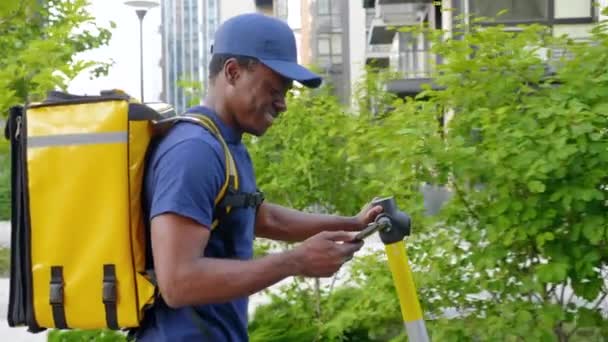 Kurir Afrika-Amerika berjalan dengan skuter listrik menggunakan smartphone menavigasi — Stok Video