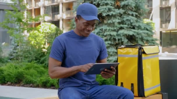 Sorrindo afro-americano homem mensageiro sentado no banco usa tablet olhando câmera — Vídeo de Stock