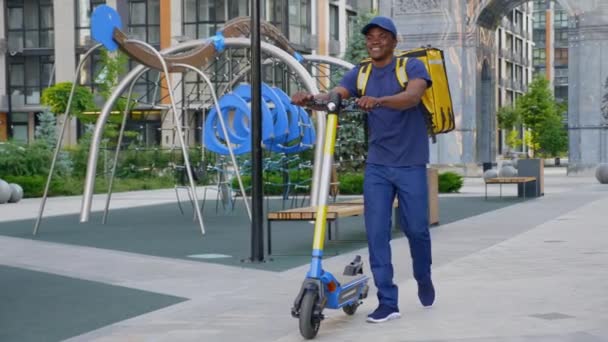 Gülümseyen Afro-Amerikan kurye teslimatı elektrikli scooterla caddede yürüyor — Stok video