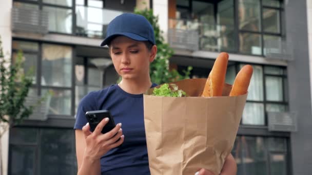 Comida entrega mensajero mujer utiliza teléfono inteligente sostiene bolsa de papel buscando dirección — Vídeos de Stock