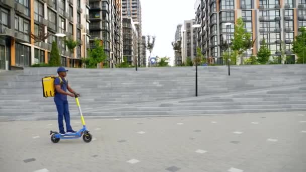 Afro-amerikai férfi futár szállítás hátizsákkal lovagol utcai elektromos robogó — Stock videók