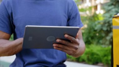 Afro-Amerikan kurye yemek teslimatını kapatın. Bankta otururken tablet kullanıyor.
