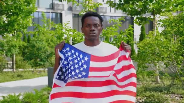 Zoom in afro-americano uomo in possesso di una bandiera americana, guarda la fotocamera e dice USA — Video Stock
