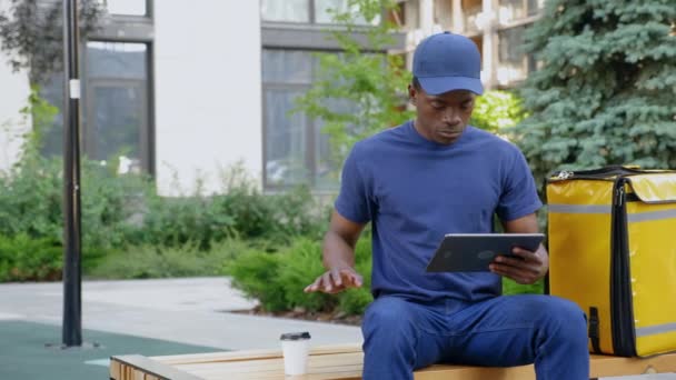 Homem africano correio entrega de alimentos sentado no banco usa tablet beber café — Vídeo de Stock