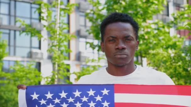 Acercarse hombre afroamericano sosteniendo una bandera americana y mira la cámara — Vídeos de Stock