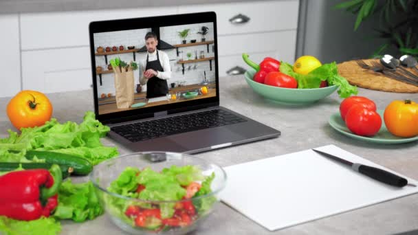 Laptop με man chef στην οθόνη στέκεται στο τραπέζι της κουζίνας κοντά σε λαχανικά τροφίμων — Αρχείο Βίντεο