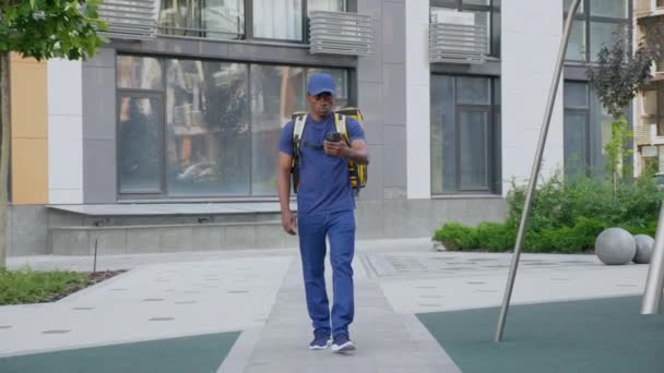 Αφρικανικός άνθρωπος courier παράδοση με σακίδιο βόλτες δρόμο βλέποντας στο smartphone — Αρχείο Βίντεο