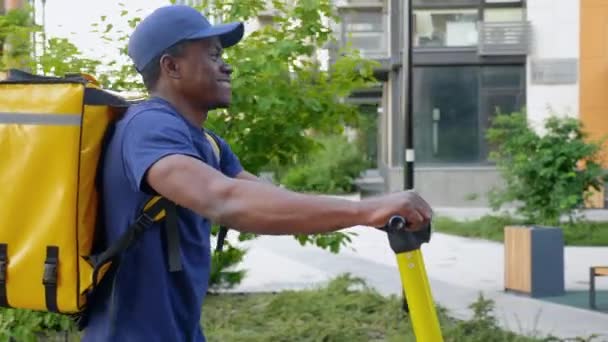 Усміхнений афроамериканський чоловік кур'єрська доставка йде вулицею з електричним скутером — стокове відео