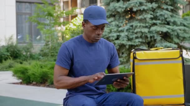 Увеличение масштаба камеры афро-американец курьер доставки сидя на скамейке использует планшет — стоковое видео