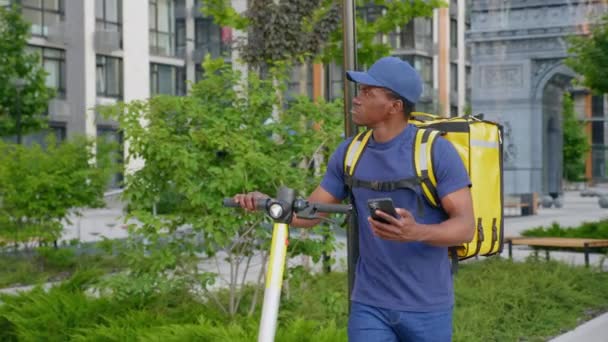 Afro-américain homme courrier promenades avec scooter électrique utilise smartphone naviguer — Video