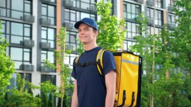 Teslimatçı pozitiftir. Arka plandaki modern binada tişört ve şapka giyer.