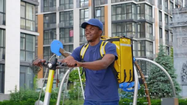 Sonriente hombre afro-americano mensajero entrega camina calle con scooter eléctrico — Vídeos de Stock