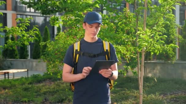 Ampliar cámara en mensajero joven entrega de comida con mochila térmica utiliza tablet — Vídeos de Stock