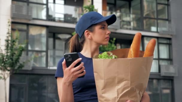 Comida entrega mensajero mujer utiliza teléfono inteligente sostiene bolsa de papel buscando dirección — Vídeo de stock