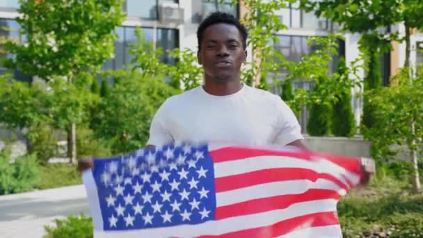 Sonriente hombre afro-americano sosteniendo una bandera americana, mira a cámara y dice Estados Unidos — Vídeo de stock