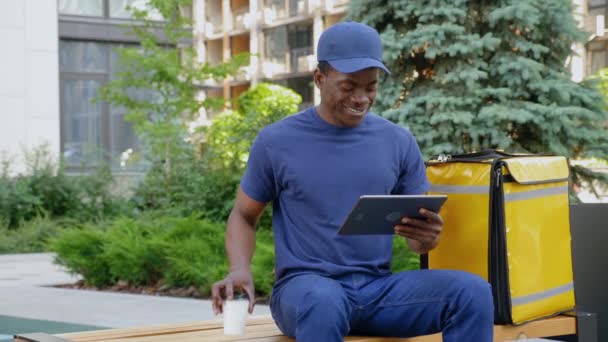 Sorridente uomo africano corriere seduto sulla panchina utilizza tablet bere caffè o tè — Video Stock