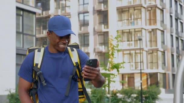 Sorrindo homem africano correio entrega de alimentos caminha rua assistindo no smartphone — Vídeo de Stock