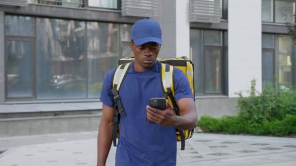 Entrega de mensajería hombre africano con mochila camina calle viendo en smartphone — Vídeo de stock