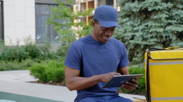 Gülümseyen Afro-Amerikan kurye yemek teslimatı bankta otururken tablet kullanıyor — Stok video