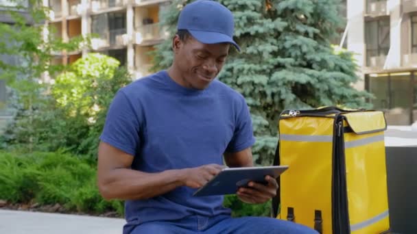 Sorridente afro-americano uomo corriere seduto sulla panchina utilizza tablet cercando fotocamera — Video Stock