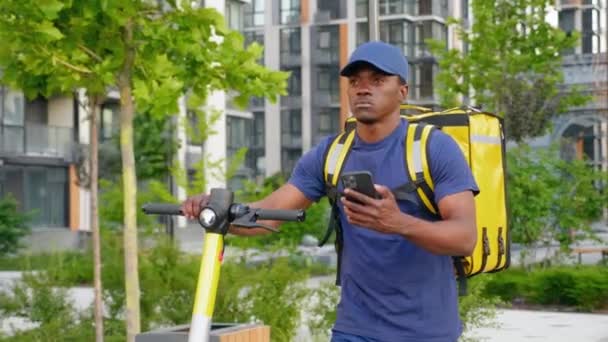 Afro-américain homme courrier promenades avec scooter électrique utilise smartphone naviguer — Video