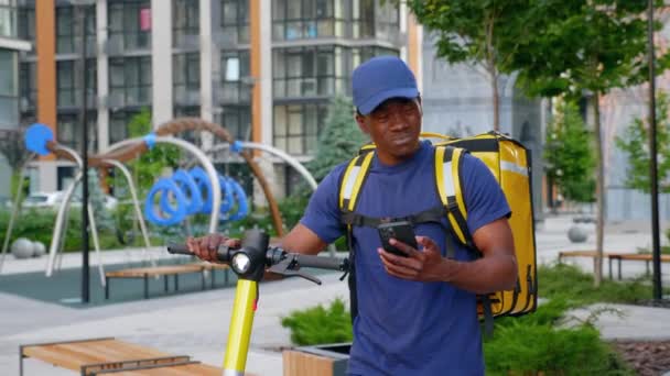 Afro-americano uomo corriere passeggiate con scooter elettrico utilizza smartphone navigare — Video Stock