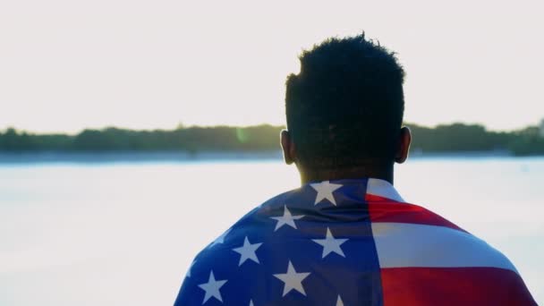 Un homme afro-américain avec le drapeau américain sur les épaules regarde loin au lever du soleil — Video
