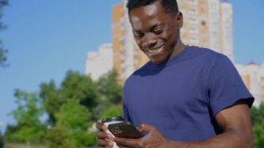 Gülümseyen Afro-Amerikalı adam sokakta yürürken cep telefonu bardağı ve kahve kullanıyor.