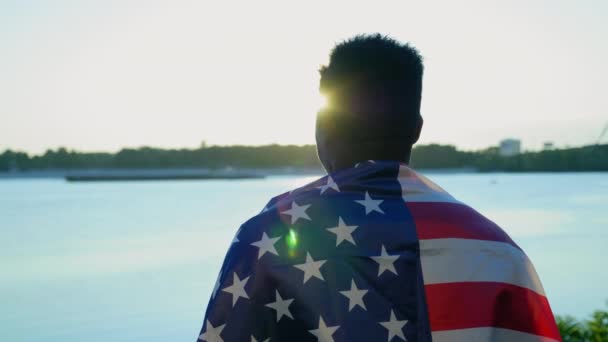 Hombre afroamericano con bandera americana sobre hombros mira a la distancia al amanecer — Vídeo de stock
