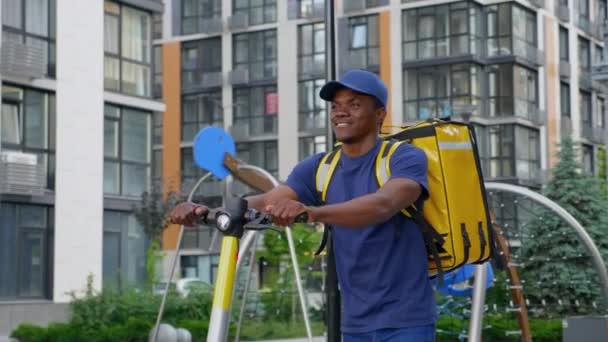 Smiling afro-american man courier delivery walks street with electric scooter — Stock Video