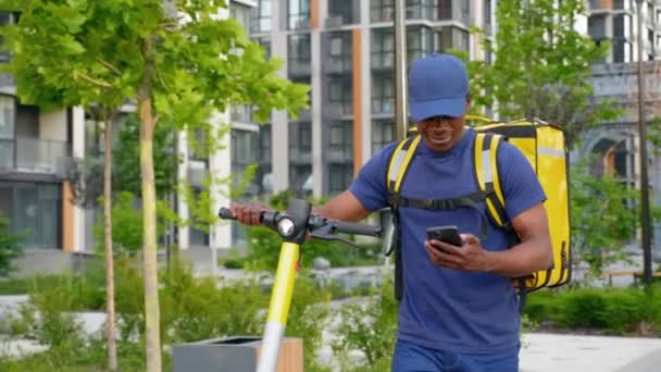 Homem afro-americano mensageiro caminha com scooter elétrico usa smartphone navegar — Vídeo de Stock