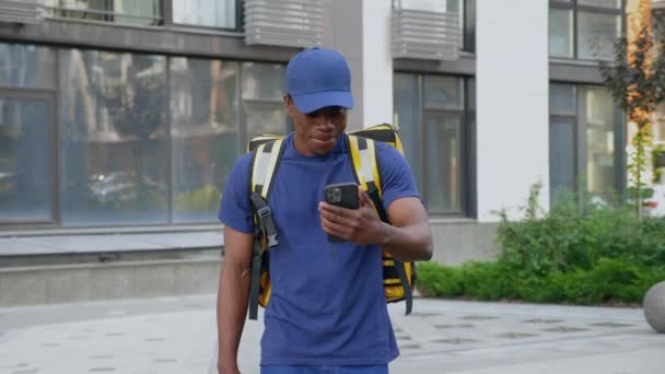 Sonriente hombre africano mensajero comida entrega paseos calle viendo en smartphone — Vídeos de Stock