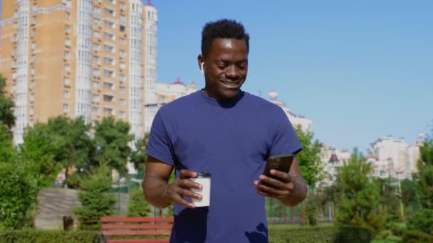 Afro-Amerikaanse zakenman in draadloze oortelefoons maakt gebruik van smartphone drinken koffie — Stockvideo