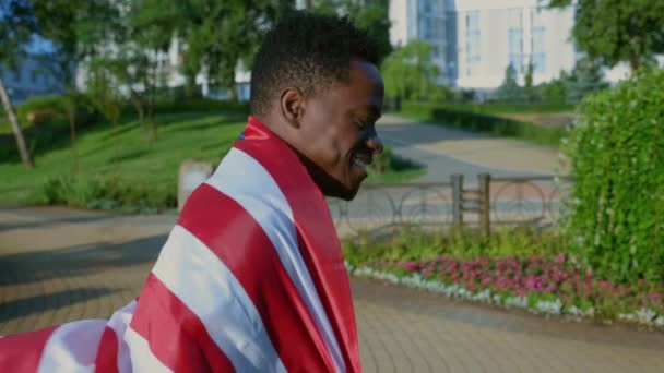 Πλευρική άποψη χαμογελώντας αφρο-Αμερικανός άνθρωπος περπάτημα δρόμο με αμερικανική σημαία στην πλάτη — Αρχείο Βίντεο