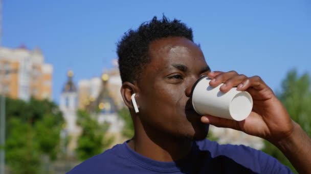 Close-up glimlachend afro-amerikaanse man het dragen van draadloze oortelefoons drinkt koffie thee — Stockvideo