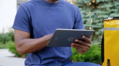 Afro-Amerikan kurye yemek teslimatını kapatın. Bankta otururken tablet kullanıyor.