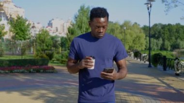 Afro-Amerikalı adam elinde kahve ya da çay olan beyaz bardakla cep telefonu kullanıyor.
