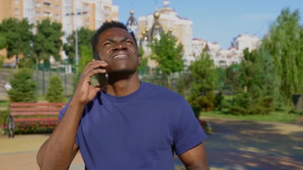 Афро-американец, стоящий в парке и разговаривающий по мобильному телефону и оглядывающийся вокруг — стоковое видео