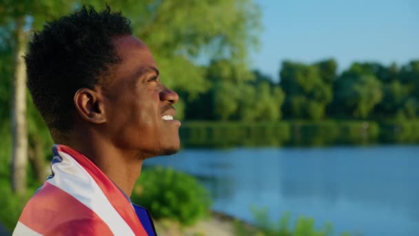 Sonriente hombre afroamericano con bandera americana en los hombros mira al amanecer — Vídeos de Stock