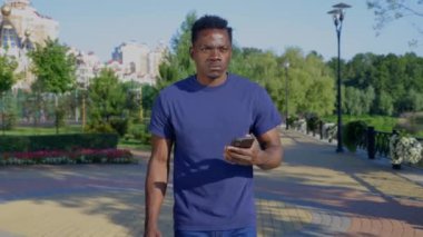 Afro-Amerikalı adam parkta yürürken akıllı telefondan navigasyona bakıyor.